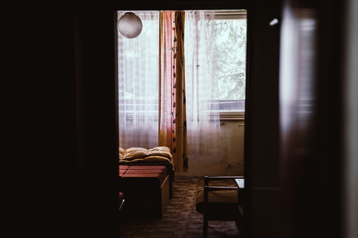 A view through a doorway into a plain guest room: a narrow bed with folded bedding, a lace curtain against a bright window, and the room itself in half-darkness