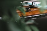A vintage car interior with rich brown leather seats is partially obscured by green leaves, suggesting a natural setting. The reflections on the car windows and polished surfaces enhance the luxurious feel of the vehicle.