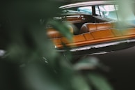 A vintage car interior with rich brown leather seats is partially obscured by green leaves, suggesting a natural setting. The reflections on the car windows and polished surfaces enhance the luxurious feel of the vehicle.