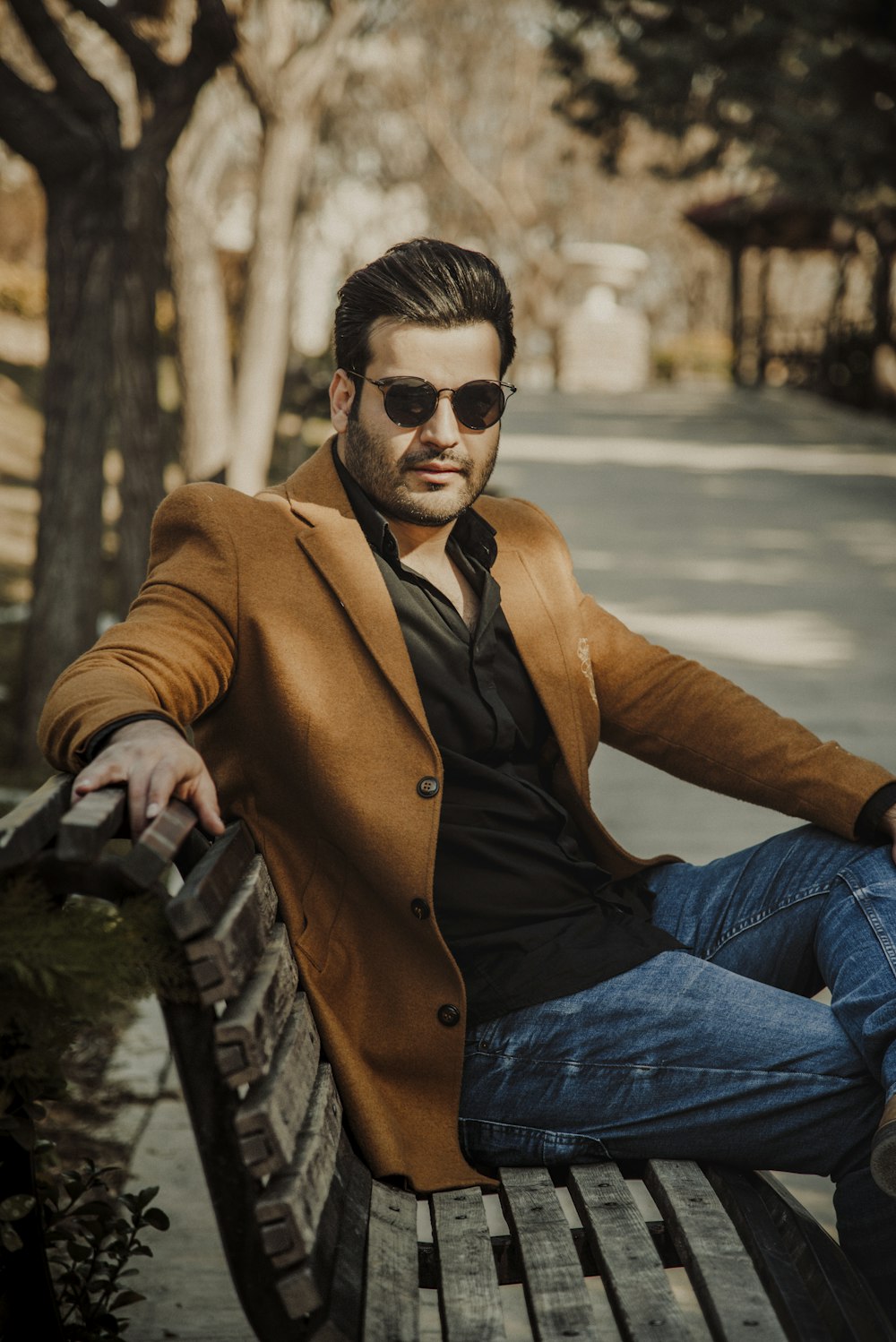 man in brown coat and blue denim jeans wearing black sunglasses sitting on black bench