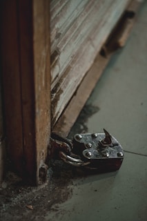 A large, weathered padlock is securely fastened to a metal latch on a rusty, corrugated metal surface. The padlock's body is dark with visible metal rivets and a looped shackle. The surrounding area appears dusty and the metal surfaces show signs of wear and oxidation.