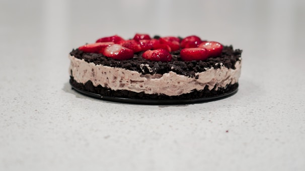 A round dessert placed on a light-colored surface features a creamy layer interspersed with dark chocolate crumb crust and is topped with sliced red strawberries.