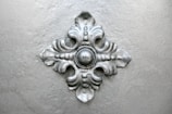 A metallic, decorative ornate piece with a symmetrical design featuring a central round element surrounded by four elegant, curved extensions resembling leaves.