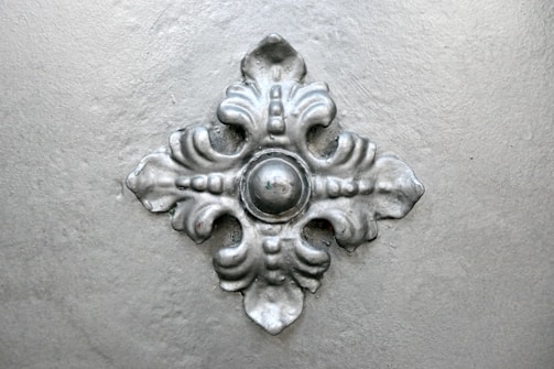 A metallic, decorative ornate piece with a symmetrical design featuring a central round element surrounded by four elegant, curved extensions resembling leaves.