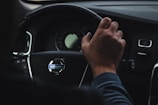 A person is gripping the steering wheel of a car with the Volvo logo clearly visible in the center. The dashboard is partially visible, displaying a speedometer and other controls. The hand on the wheel appears relaxed, suggesting a calm driving environment.