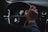 A person is gripping the steering wheel of a car with the Volvo logo clearly visible in the center. The dashboard is partially visible, displaying a speedometer and other controls. The hand on the wheel appears relaxed, suggesting a calm driving environment.