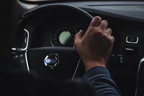 A person is gripping the steering wheel of a car with the Volvo logo clearly visible in the center. The dashboard is partially visible, displaying a speedometer and other controls. The hand on the wheel appears relaxed, suggesting a calm driving environment.