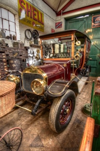 A vintage automobile is displayed in a garage with various automotive tools and memorabilia. The car features a polished maroon and gold exterior, with a wicker basket attached to the front. Surrounding the car are walls filled with old automotive signs and parts. The flooring is patterned, and there's an overall vintage and nostalgic atmosphere in the setting.