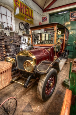 A vintage automobile is displayed in a garage with various automotive tools and memorabilia. The car features a polished maroon and gold exterior, with a wicker basket attached to the front. Surrounding the car are walls filled with old automotive signs and parts. The flooring is patterned, and there's an overall vintage and nostalgic atmosphere in the setting.