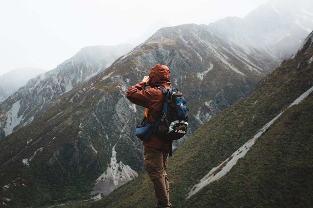 man in brown jacket and black backpack standing on rock mountain during daytime