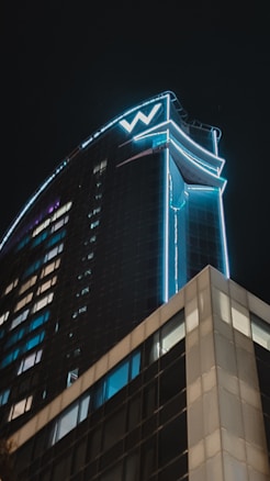 A tall, modern skyscraper is illuminated against the night sky, featuring a prominent 'W' logo at the top. The building has a sleek, curved design with bright blue neon lights outlining its edges and sections of its façade. Windows are arranged in a grid pattern, reflecting the lights cityscape.