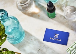 A neatly arranged layout on a marble surface features a turquoise glass bottle, a dropper bottle labeled 'Rejuvia', and two beakers with measurements. A blue business card with the Rejuvia logo and a nearby green and white patterned box add a pop of color. Fresh green leaves are partially visible in the bottom left corner, suggesting a natural theme.