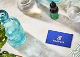 A neatly arranged layout on a marble surface features a turquoise glass bottle, a dropper bottle labeled 'Rejuvia', and two beakers with measurements. A blue business card with the Rejuvia logo and a nearby green and white patterned box add a pop of color. Fresh green leaves are partially visible in the bottom left corner, suggesting a natural theme.