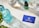 A neatly arranged layout on a marble surface features a turquoise glass bottle, a dropper bottle labeled 'Rejuvia', and two beakers with measurements. A blue business card with the Rejuvia logo and a nearby green and white patterned box add a pop of color. Fresh green leaves are partially visible in the bottom left corner, suggesting a natural theme.