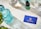 A neatly arranged layout on a marble surface features a turquoise glass bottle, a dropper bottle labeled 'Rejuvia', and two beakers with measurements. A blue business card with the Rejuvia logo and a nearby green and white patterned box add a pop of color. Fresh green leaves are partially visible in the bottom left corner, suggesting a natural theme.