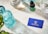 A neatly arranged layout on a marble surface features a turquoise glass bottle, a dropper bottle labeled 'Rejuvia', and two beakers with measurements. A blue business card with the Rejuvia logo and a nearby green and white patterned box add a pop of color. Fresh green leaves are partially visible in the bottom left corner, suggesting a natural theme.