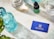 A neatly arranged layout on a marble surface features a turquoise glass bottle, a dropper bottle labeled 'Rejuvia', and two beakers with measurements. A blue business card with the Rejuvia logo and a nearby green and white patterned box add a pop of color. Fresh green leaves are partially visible in the bottom left corner, suggesting a natural theme.