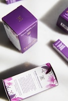 Packages of premium hemp softgels are arranged on a surface. The boxes are predominantly purple with white panels and floral designs. The front of the packaging displays the product name, 'Pure Tranquility', and a brief description of the benefits.