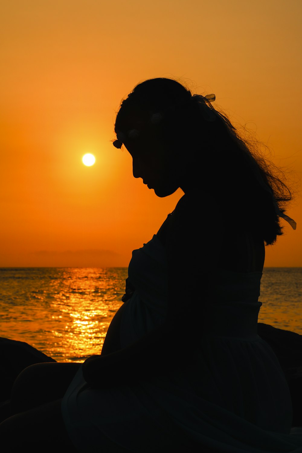 Silhouette einer Frau, die während des Sonnenuntergangs auf einem Felsen in der Nähe eines Gewässers sitzt