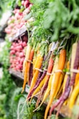 A vibrant display of fresh produce in a market or grocery setting. Bundles of colorful carrots, featuring hues of orange, yellow, and purple, are neatly arranged alongside other vegetables. Green leafy tops of the carrots add a lush element to the scene, enhancing the freshness. In the background, more vegetables like radishes contribute to the overall assortment.