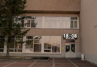 A building facade with large windows and a digital clock displaying the time 18:08. The front entrance features glass doors, and a tree is visible to the left. The setting is subdued, with minimal activity noticeable.