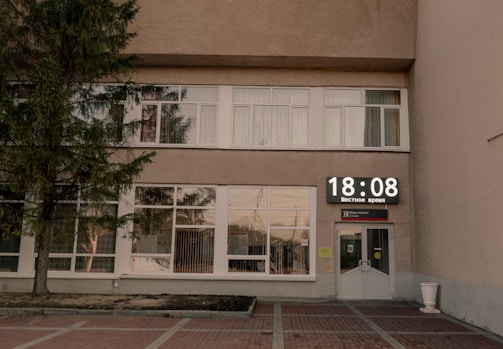 A building facade with large windows and a digital clock displaying the time 18:08. The front entrance features glass doors, and a tree is visible to the left. The setting is subdued, with minimal activity noticeable.