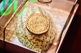 A delicately crafted dessert presented in a clear box tied with a golden string. It features a decorative piece resembling a coin with the text 'Cakeerie Vitoria' embossed on it. The dessert itself appears to be speckled, possibly with seeds or chocolate chips.
