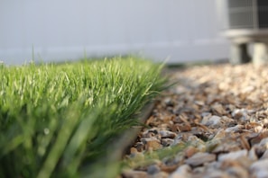 A close-up perspective of a neatly cut grassy lawn bordered by a line of small, multicolored stones. In the background, an out-of-focus structure or unit suggests a domestic outdoor setting.