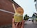 A hand holds a cup of ice cream with a biscuit on top, featuring the text 'Vanhan Porvoon Jäätelotehdas' on the cup and biscuit. The background shows a cobblestone street lined with buildings, a few trees, and a white tent, suggesting a market or street event.