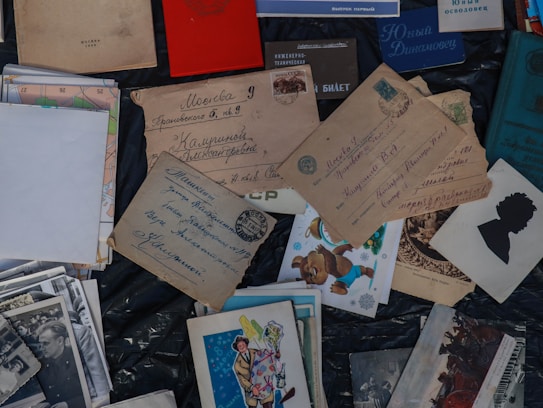 A collection of vintage postcards and envelopes with handwritten addresses is spread out. There are also old photographs and children's illustration cards among the items. Some of the envelopes have vintage stamps affixed. Russian text is visible on several of the items.