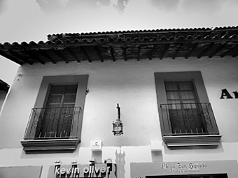 A black and white photograph of a building with two windows that have decorative wrought iron railings. The building has a slightly textured surface and a sloped, tiled roof with exposed wooden beams beneath. A lantern is mounted between the windows, above a sign reading 'Plaza San Hipolito' with smaller text 'Plata y artesan&iacute;as.'