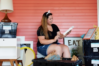 A woman is sitting in front of a coral-colored wall, surrounded by various moving boxes and home decor items. She is holding a picture frame and appears to be smiling. There is a black plastic bin labeled 'HIVE BOXX' and other decorative items like a lamp and framed art pieces around her.