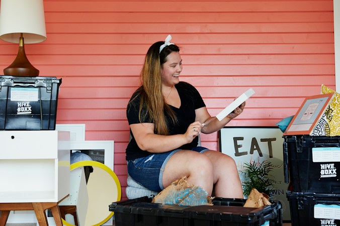 A woman is sitting in front of a coral-colored wall, surrounded by various moving boxes and home decor items. She is holding a picture frame and appears to be smiling. There is a black plastic bin labeled 'HIVE BOXX' and other decorative items like a lamp and framed art pieces around her.