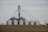 A cluster of large silver grain silos stands on a barren field, connected by a network of metal piping. The sky is overcast, contributing to a muted atmosphere. The structures are cylindrical with domed roofs, typical of agricultural storage facilities.