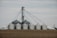 A cluster of large silver grain silos stands on a barren field, connected by a network of metal piping. The sky is overcast, contributing to a muted atmosphere. The structures are cylindrical with domed roofs, typical of agricultural storage facilities.
