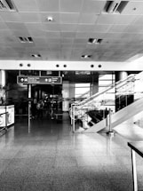 A modern airport interior with a wide hallway featuring shiny tiled floors, a high ceiling with embedded lights, and directional signs overhead pointing towards baggage claim and transfer. To the right, there is an escalator with glass railings. A few people are walking towards the sign.