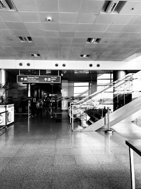 A modern airport interior with a wide hallway featuring shiny tiled floors, a high ceiling with embedded lights, and directional signs overhead pointing towards baggage claim and transfer. To the right, there is an escalator with glass railings. A few people are walking towards the sign.
