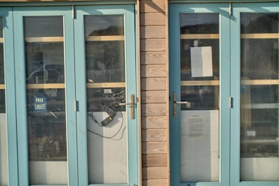 A set of blue double glass doors set in a wooden frame. One door has a small sign with the words 'Free Sauces' taped inside. There are some cables and an electronic device visible through the glass. Another paper notice is attached to the door, containing text about COVID-19 guidelines.