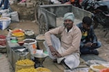 An outdoor market scene with a vendor sitting cross-legged on the ground near a cart, surrounded by bags of various colorful spices and goods. A man is sitting next to him using a mobile phone. Motorcycles are parked in the background.