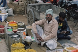 An outdoor market scene with a vendor sitting cross-legged on the ground near a cart, surrounded by bags of various colorful spices and goods. A man is sitting next to him using a mobile phone. Motorcycles are parked in the background.