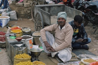 An outdoor market scene with a vendor sitting cross-legged on the ground near a cart, surrounded by bags of various colorful spices and goods. A man is sitting next to him using a mobile phone. Motorcycles are parked in the background.