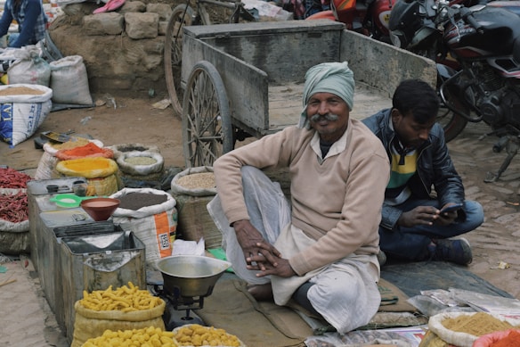 An outdoor market scene with a vendor sitting cross-legged on the ground near a cart, surrounded by bags of various colorful spices and goods. A man is sitting next to him using a mobile phone. Motorcycles are parked in the background.