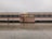 A small, light-colored utility vehicle is positioned in front of a large passenger train. The train features mostly gray and red tones and stands on a wet platform reflected in the rain-soaked pavement. The sky appears overcast, suggesting a gray and gloomy setting.