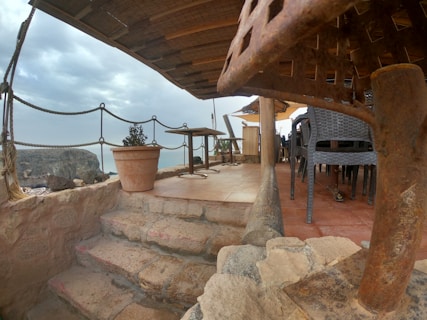 A rustic outdoor seating area with stone steps leading up to a partially covered terrace. The area is furnished with wicker chairs and a wooden table, and is adorned with a potted plant. Overhead, a thatched roof provides some shelter, while the view shows a cloudy sky and a distant rocky landscape.