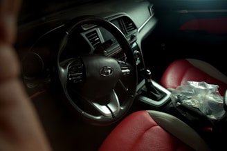 A view of a car interior featuring a steering wheel with the Hyundai logo. The gear shift is visible with a plastic bag placed nearby. The seats are upholstered in a combination of red and beige leather. The dashboard is equipped with various controls and displays.