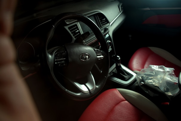 A view of a car interior featuring a steering wheel with the Hyundai logo. The gear shift is visible with a plastic bag placed nearby. The seats are upholstered in a combination of red and beige leather. The dashboard is equipped with various controls and displays.
