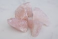 Several raw rose quartz crystals are arranged on a light surface. The stones are light pink in color with translucent qualities and have rough, jagged edges.