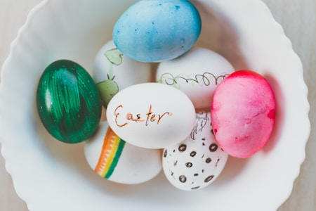 A collection of six colorful Easter eggs rests in a white, round bowl. Each egg is uniquely decorated with varying colors and patterns, including stripes, polka dots, and abstract designs. The word 'Easter' is written on one of the eggs in orange.