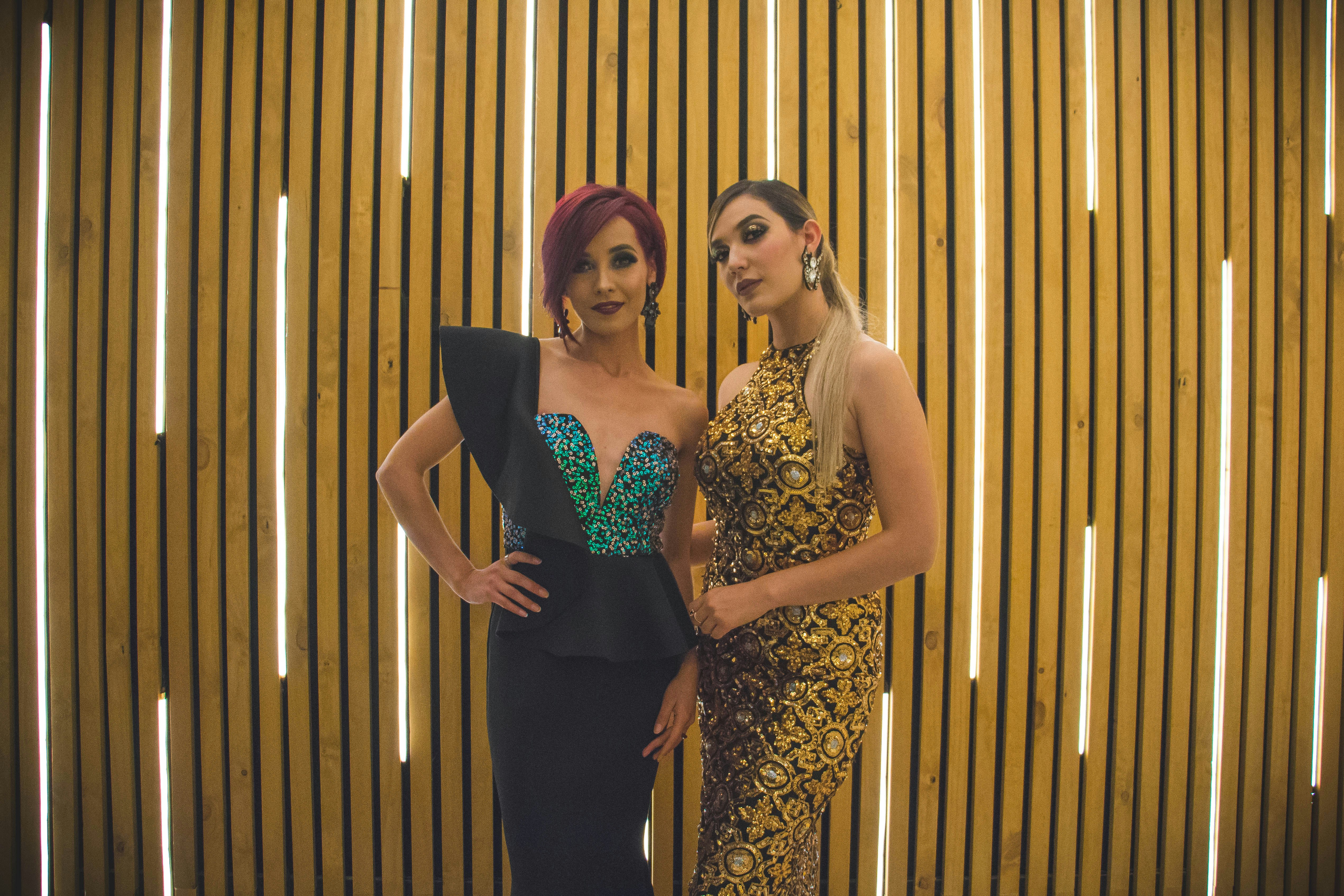 2 women in black and brown floral dresses standing beside brown wooden wall