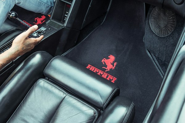 The image displays the interior of a car, highlighting the black leather upholstery and a floor mat with the iconic Ferrari logo in red. A person's hand is visible, operating the gear shift, suggesting the car's manual transmission system.